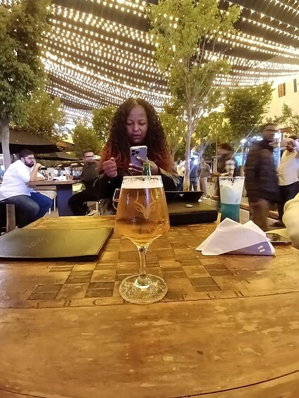 A person taking a selfie with a glass of beer on a wooden table at an outdoor cafe. 5