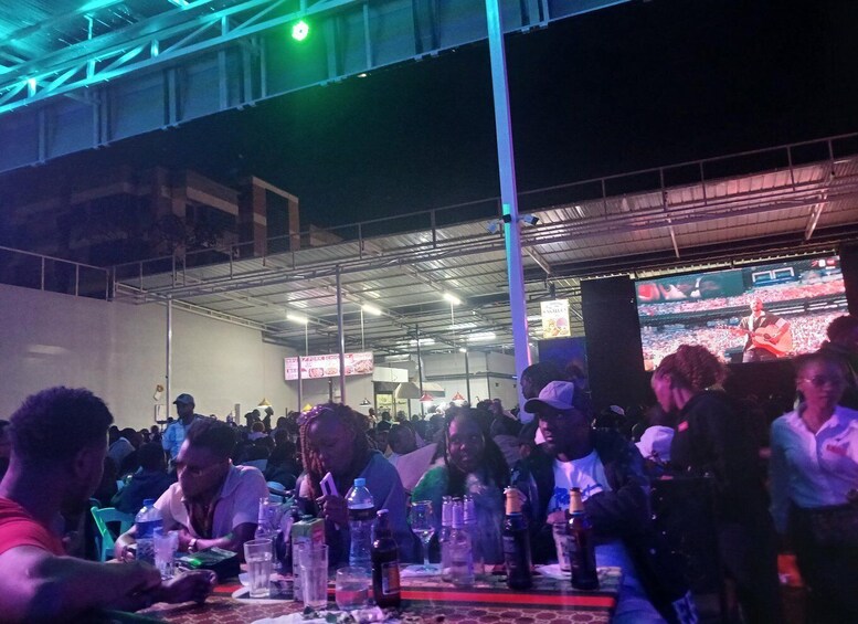A crowded indoor event with people seated at tables, a stage with a performer, and a large screen displaying the event. 3