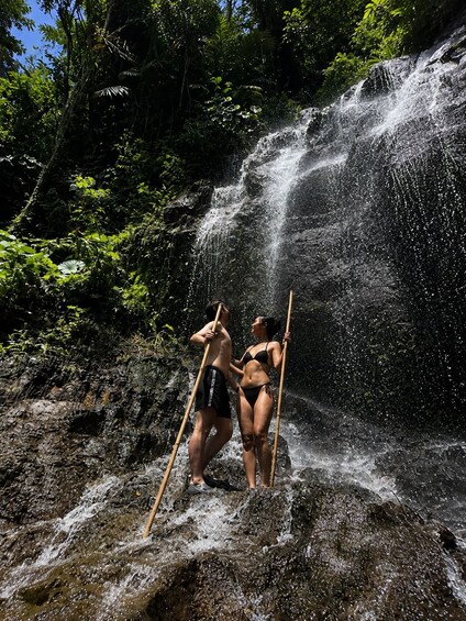 Zwei Personen stehen vor einem Wasserfall, umgeben von üppigem Grün. 3