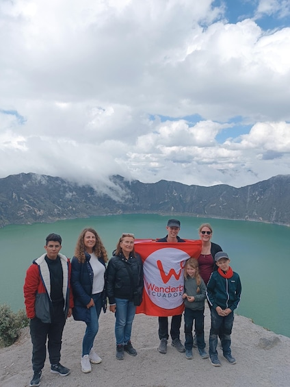 A group of people standing by a lake with mountains in the background, holding a banner that says "Wanderb Ecuador". 5