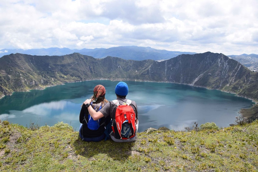 Two people sitting on a grassy hill overlooking a large lake surrounded by mountains. 1