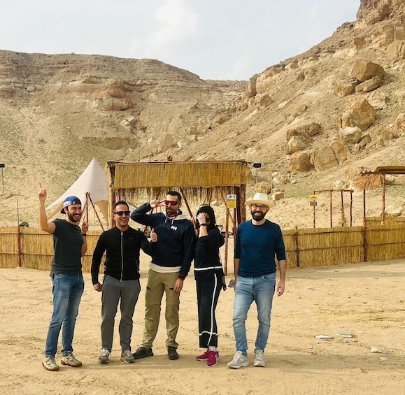 A group of people standing in a desert with rocky terrain and a thatched structure in the background. 3
