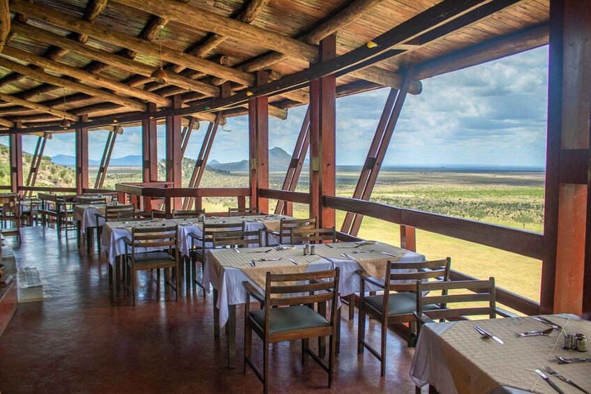 An outdoor restaurant with wooden tables and chairs, large windows, and a view of a landscape with mountains. 2