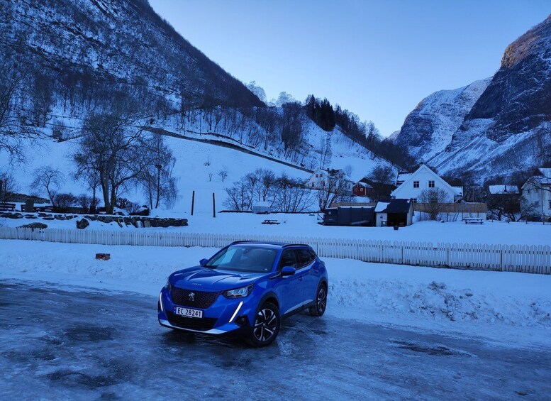A blue car parked on a snow-covered road with houses and mountains in the background. 4