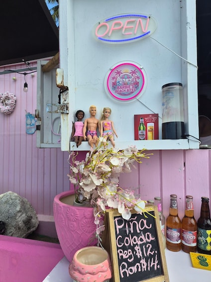 Vaaleanpunaisella teemalla sisustettu ulkotila, jossa on neonvaloin valaistu ”AVOIN”-kyltti, ”Barbie’s Beach Bites” -logo ja liitutaulusta tehty juomalista, jossa on tarjolla muun muassa ”Pina Colada” -cocktaileja. 3