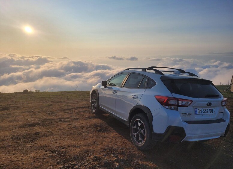 A white Subaru car parked on a dirt road with a cloudy sky and sun in the background. 1