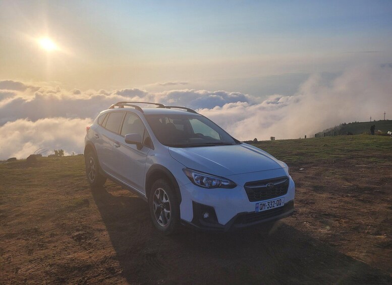 A white SUV parked on a grassy hill with clouds below and the sun in the background. 4