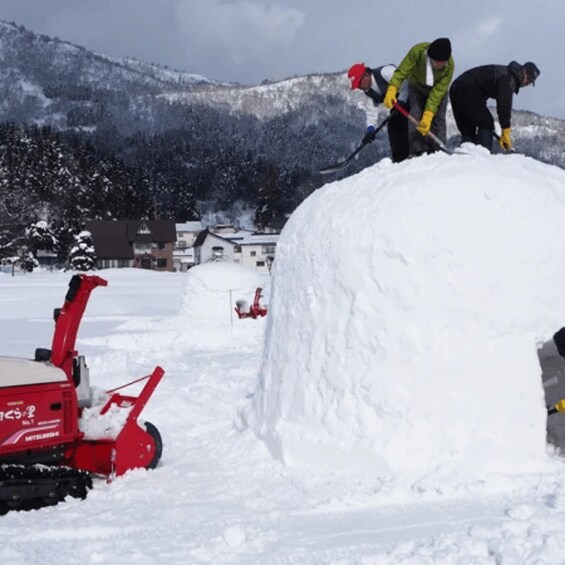 Two people shoveling snow into a large pile, with a red snow blower in the foreground. 5