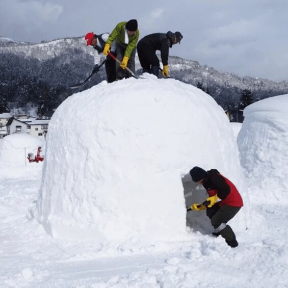 Three people building a snow structure in a snowy landscape with houses and mountains in the background. 2