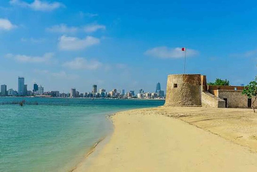 A sandy beach with a stone structure and a flag, a clear blue sky, and a city skyline in the background. 3