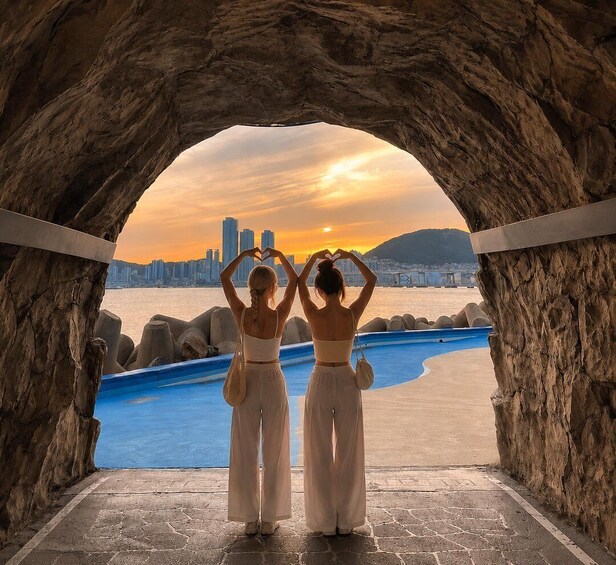 Two women making a heart shape with their hands in front of a rocky archway overlooking a body of water and a city skyline at sunset. 1
