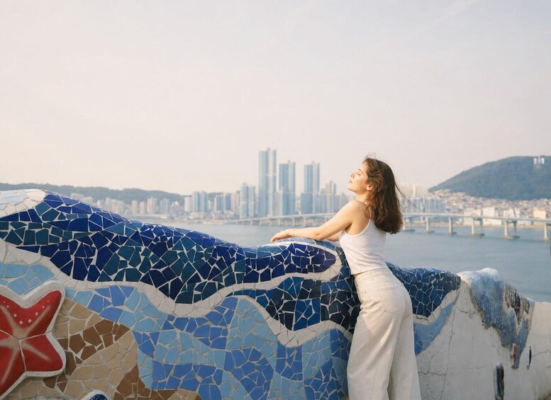 A mosaic wall with a woman leaning on it, overlooking a cityscape with a bridge and buildings. 2