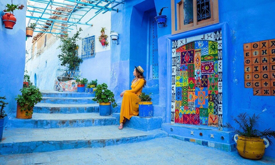 Una persona sentada en escalones azules en un callejón estrecho, con patrones geométricos de colores en las paredes y plantas en macetas. 2