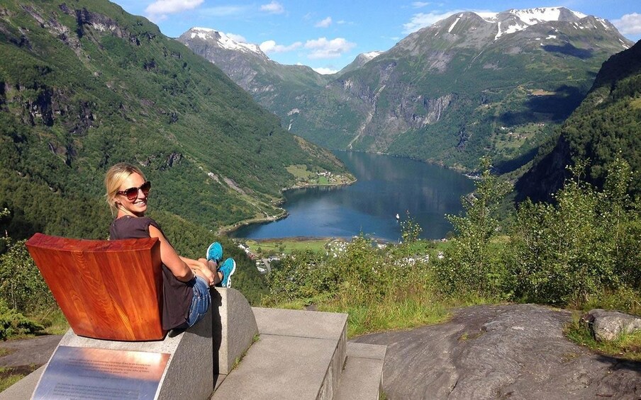 En person sitter på en träbänk med utsikt över en fjord, och i bakgrunden reser sig berg. 1