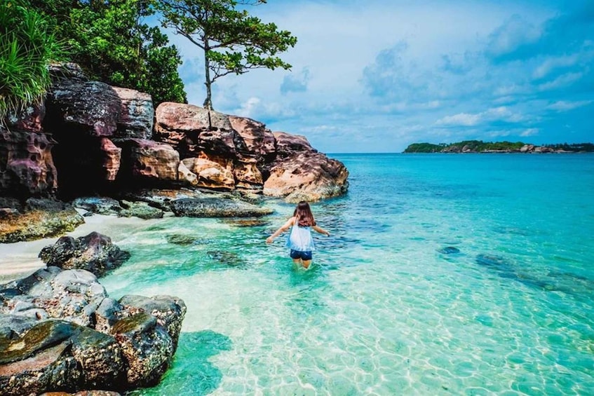 A person wading through clear turquoise water near rocky shores with a tree and distant land. 5