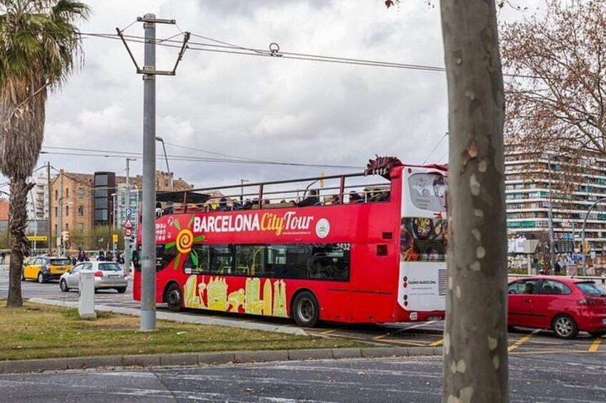 A red double-decker bus with 'BARCELONA City Tour' written on it, parked on a street with buildings and cars in the background. 3
