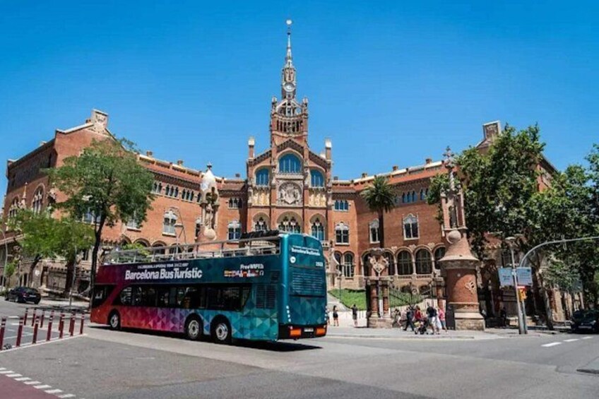 A bus with 'Barcelona Bus Kustic' written on it, a large historic building with a clock tower, and a clear blue sky. 4
