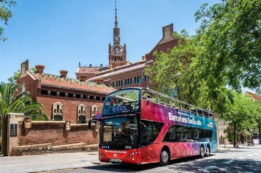 A red double-decker bus with 'Barcelona Busaric' on the side, parked in front of a historic building with a clock tower. 1