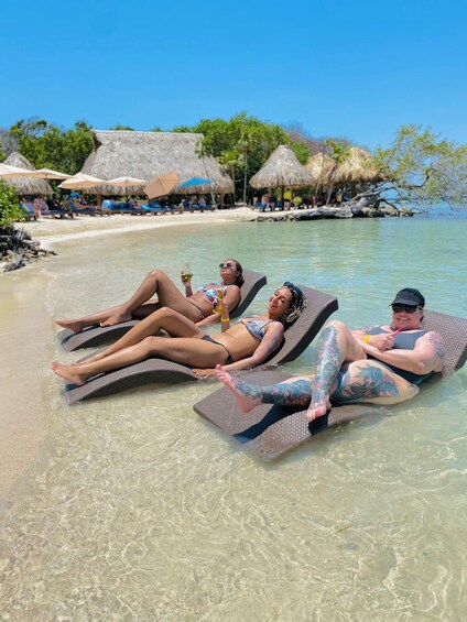 Three people sunbathing on lounge chairs by a beach with thatched umbrellas in the background. 1