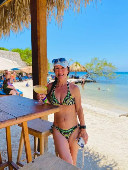 A person in a bikini holding a drink at a beachside bar with a thatched roof, palm trees, and the ocean in the background. 5