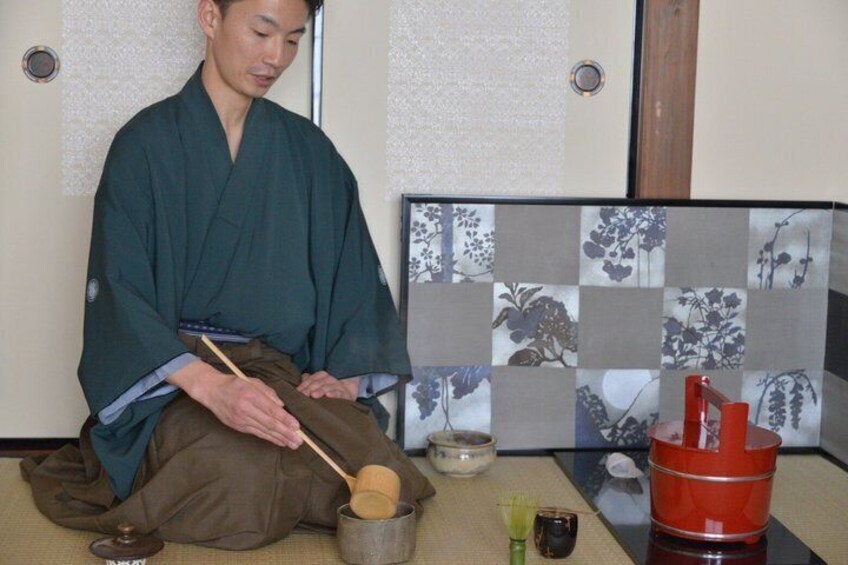 A person in traditional attire sitting on the floor, holding a wooden ladle over a bowl. 1