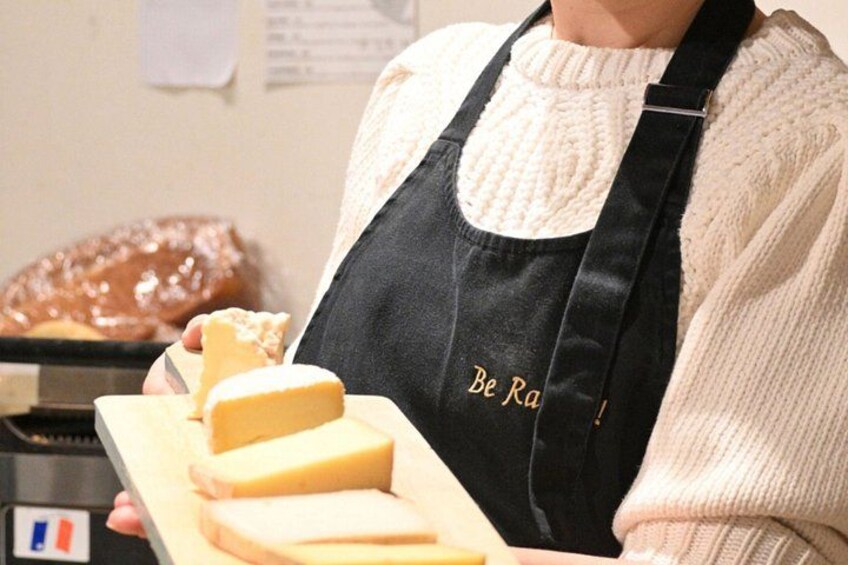 A person wearing a black apron with 'Be Ra' text, holding a wooden cutting board with cheese. 1