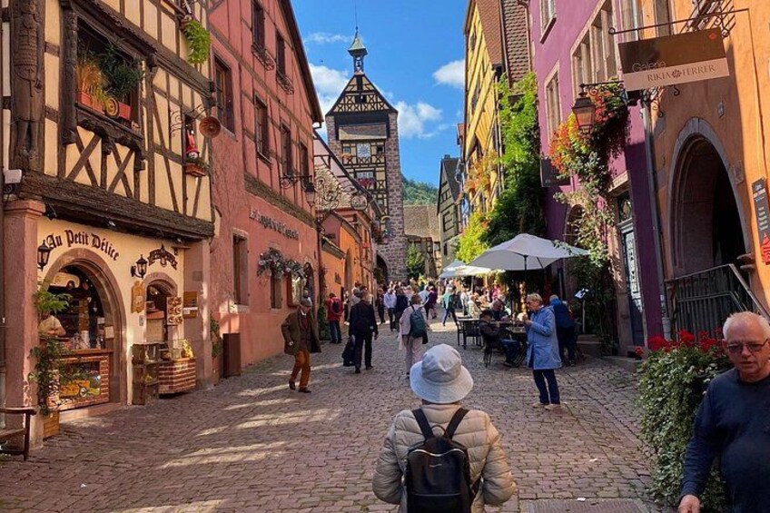 A cobblestone street lined with half-timbered buildings, a café with outdoor seating, and pedestrians walking. 2