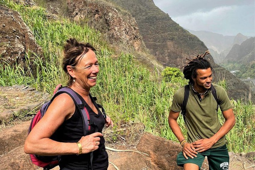 Two people hiking on a mountainous trail with greenery and rocky terrain. 5
