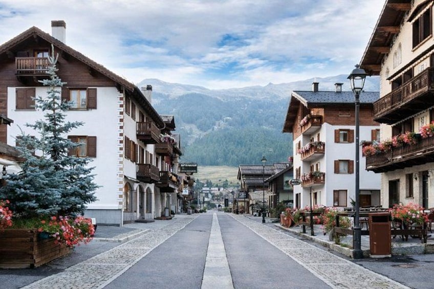 A mountain village street with wooden buildings, balconies with flowers, and a clear sky. 1