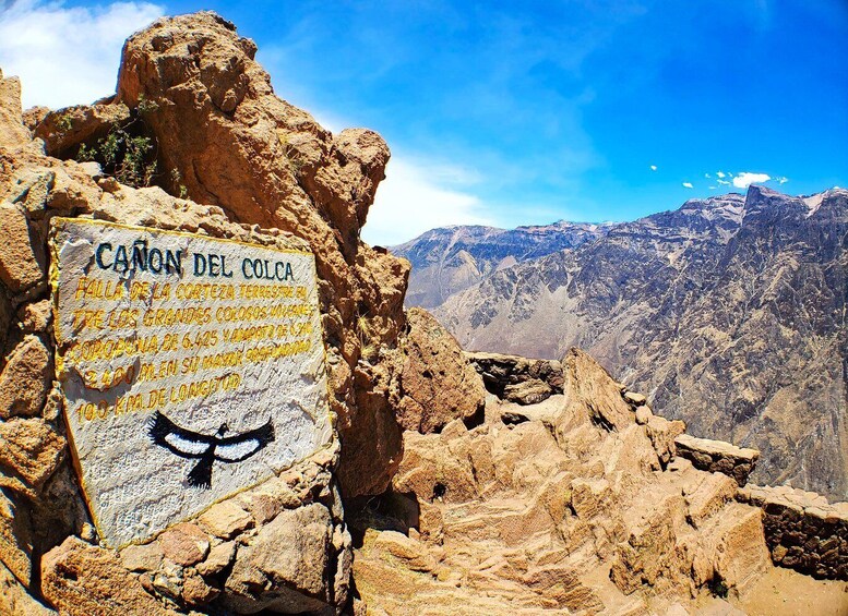A rocky terrain with a sign reading "Cañon del Colca" and a mountain range in the background. 1