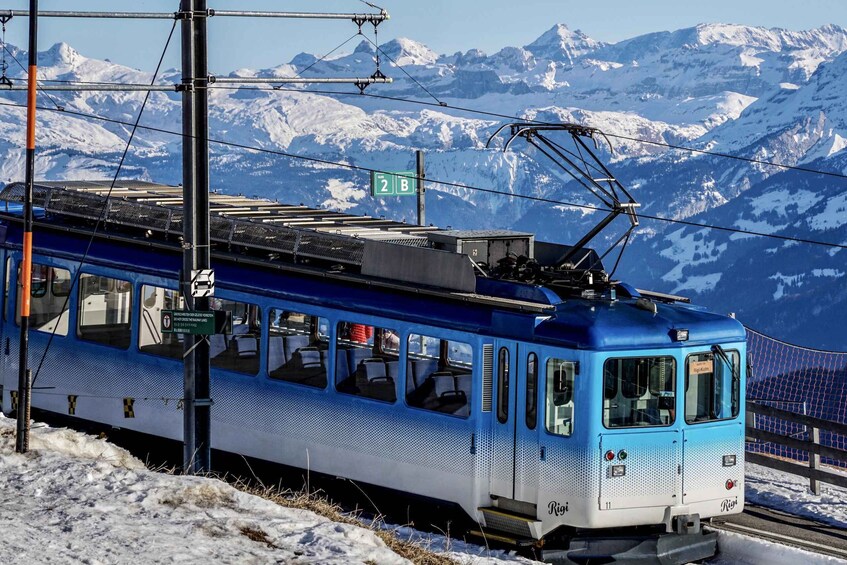 A blue and white train on tracks with snow-covered mountains in the background. 3
