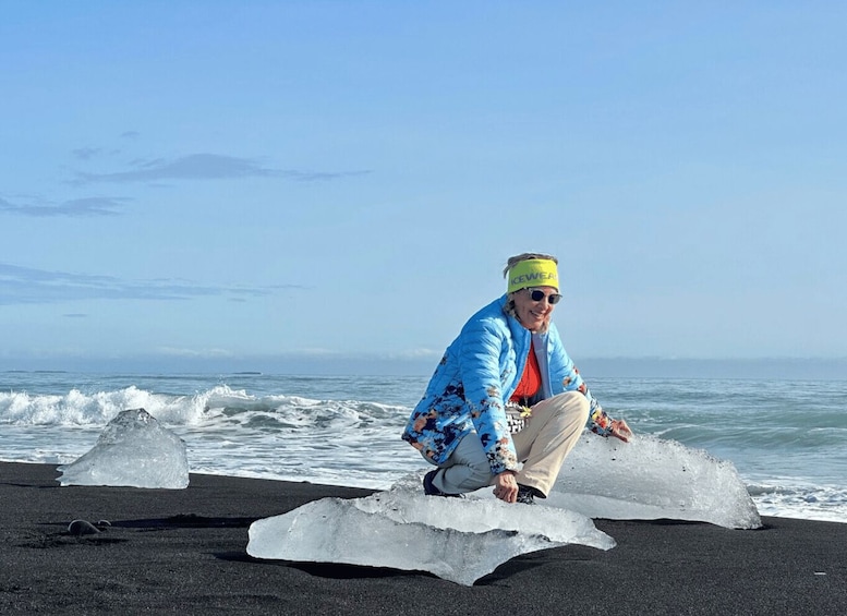 A person crouching on a black sand beach with icebergs in the water. 4