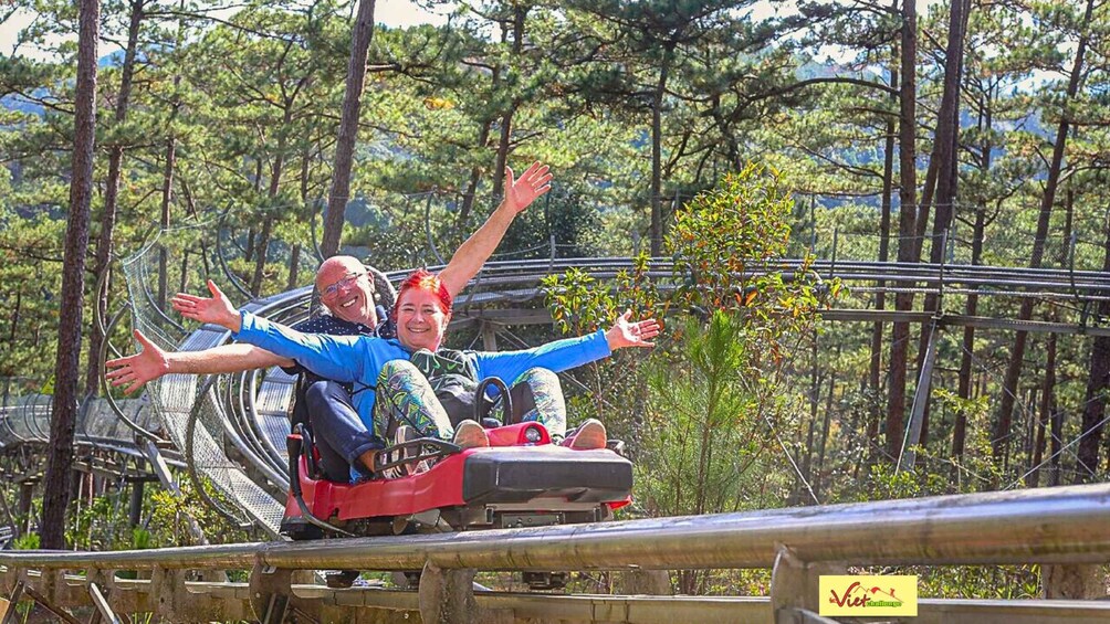 Dos personas en una montaña rusa con los brazos extendidos, rodeadas de árboles y una estructura de madera. 2