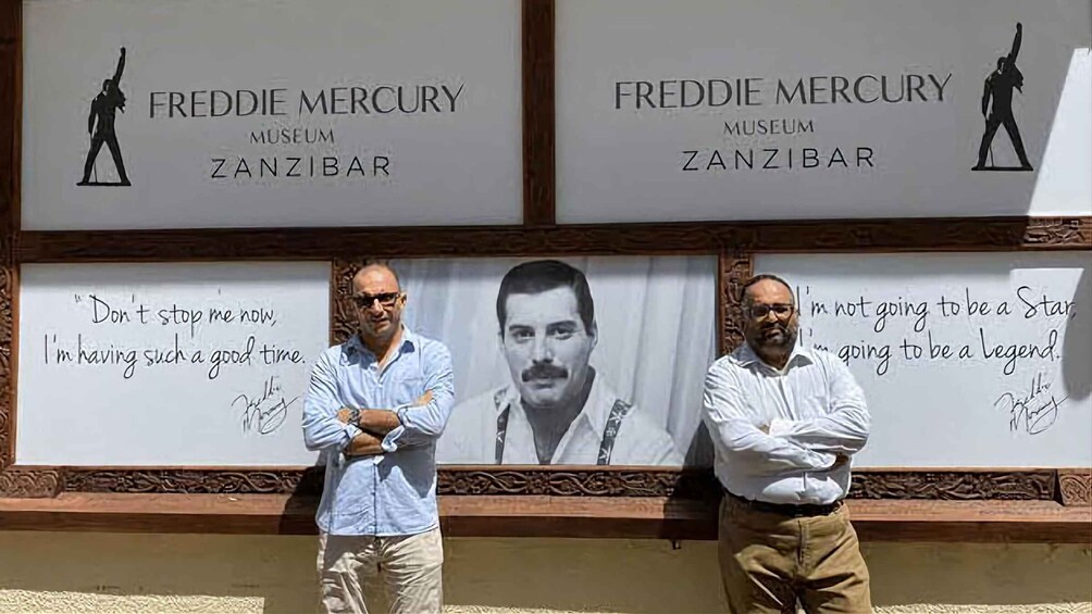 Kaksi miestä seisoo seinän edessä, ja seinässä on kylttejä, jotka osoittavat Freddie Mercuryn museoon Zanzibarissa. 3