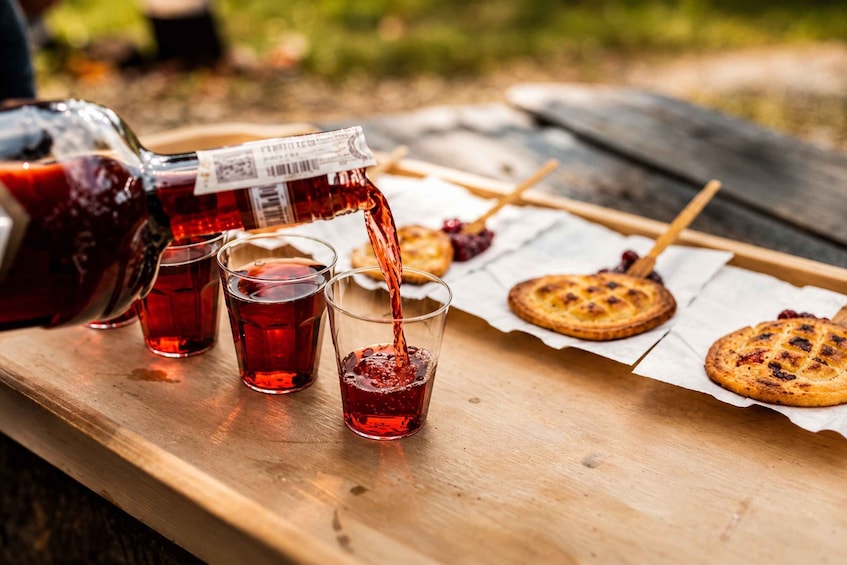 A wooden table with a bottle of red liquid being poured into glasses, cookies on a paper-lined tray, and a wooden spoon. 2