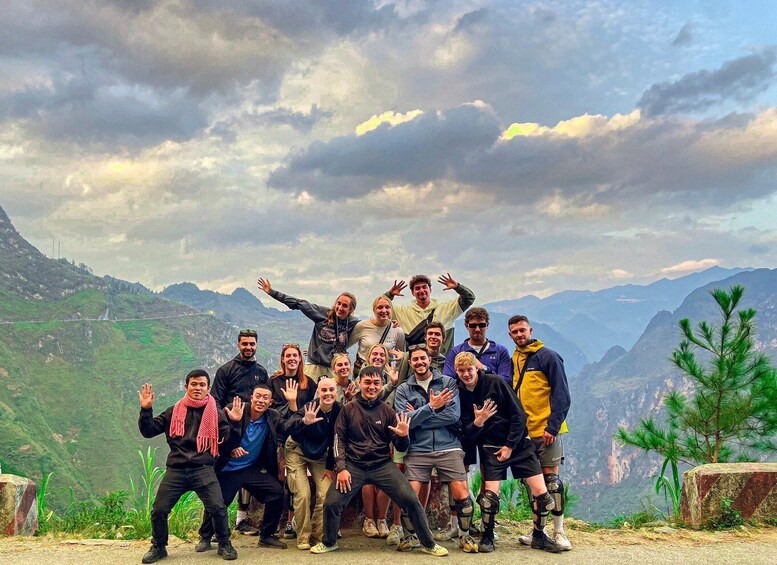 A group of people posing on a mountain ridge with a cloudy sky and green hills in the background. 1