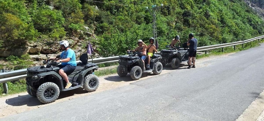 People riding ATVs on a road with a guardrail and greenery in the background. 4