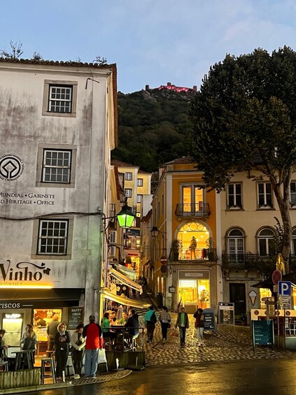 A cobblestone street with people walking, a building with 'Vinho' sign, and a hill in the background. 3