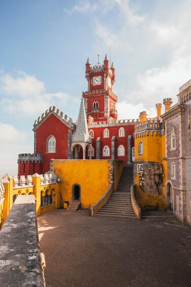 A castle with red and yellow walls, a tower with a clock, and a staircase leading to the entrance. 5