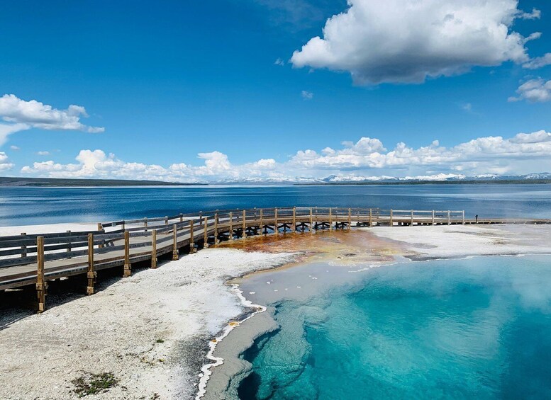 A wooden boardwalk leading to a turquoise hot spring surrounded by snow-covered mountains. 2
