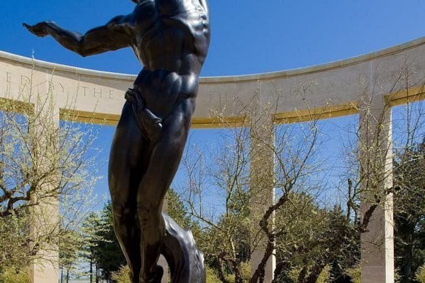 Una estatua de bronce de una figura musculosa en una pose dinámica, con un edificio de columnas y un cielo azul despejado de fondo. 1