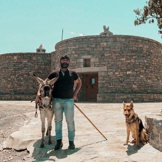 A man with a donkey and a dog in front of a stone building. 1