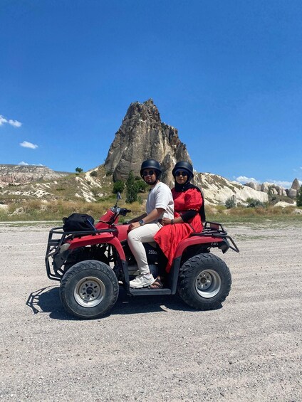 Two people on a red ATV in a rocky outdoor setting with a prominent rock formation in the background. 2