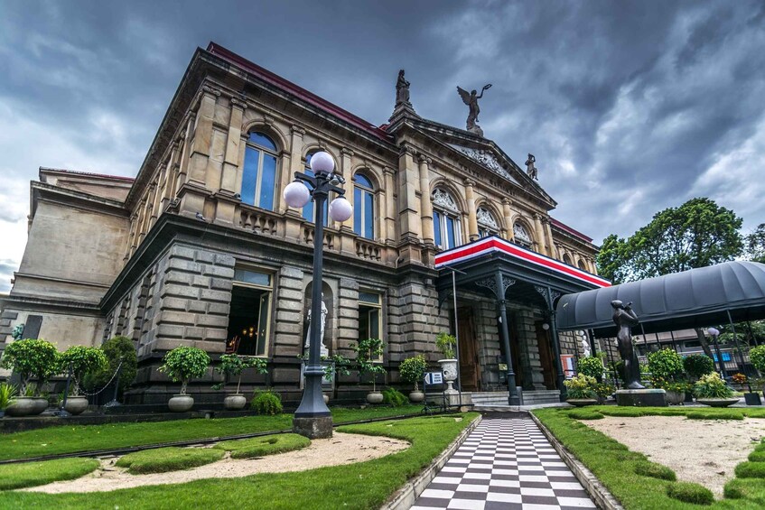 A historic building with a checkered pathway leading to the entrance, surrounded by well-maintained greenery and statues. 1