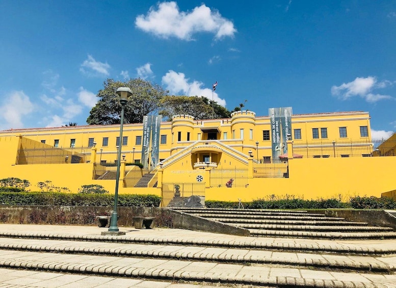 A yellow building with a staircase leading up to a balcony. There are banners and a flag on the building. 3