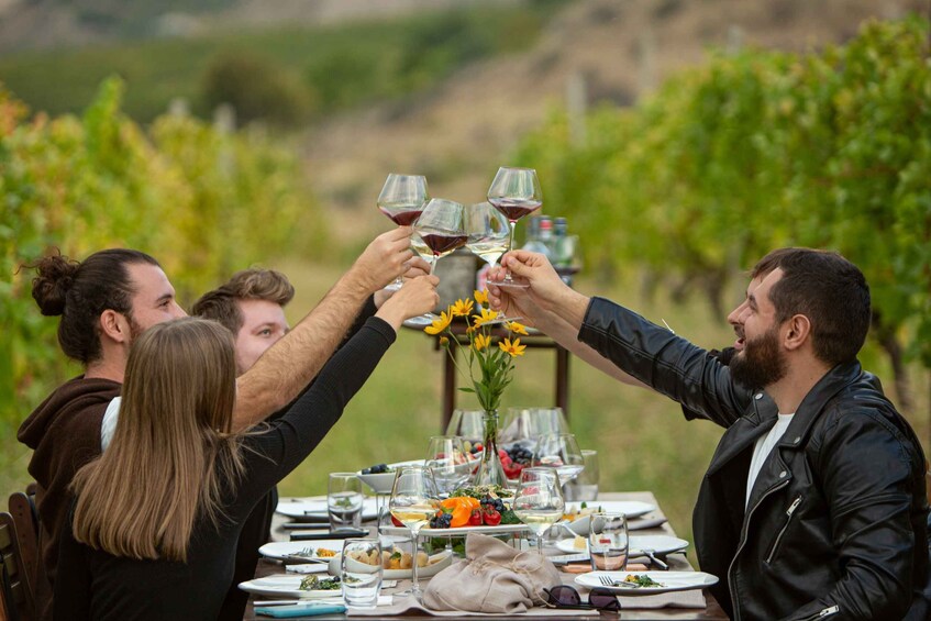 A group of people toasting with wine glasses at a vineyard table with food and flowers. 1