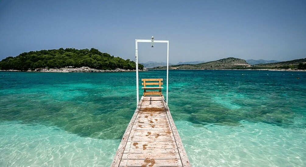 A wooden pier leading to a bench by a turquoise sea with a forested island in the background. 2