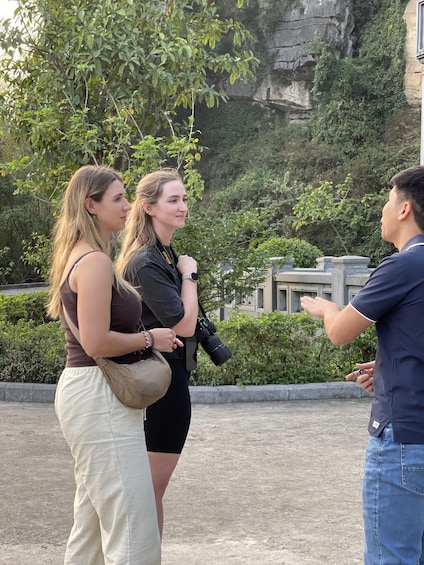 Three people standing outdoors with greenery and a building in the background. 1