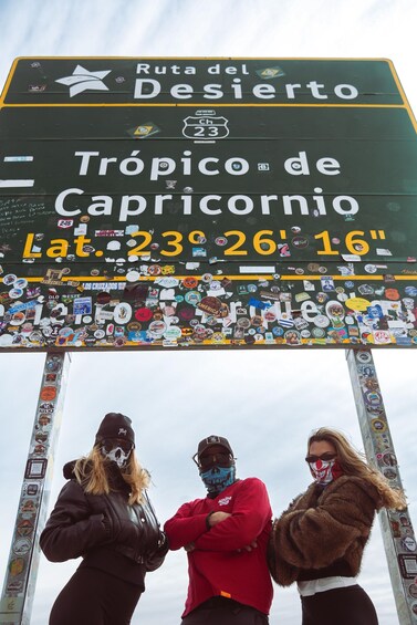 A sign with stickers, three people standing in front of it, and a cloudy sky. 2