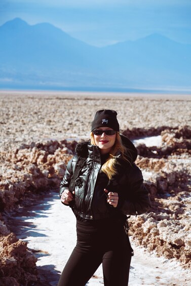 A person in a black jacket and beanie standing in a barren landscape with snow patches and mountains in the background. 4